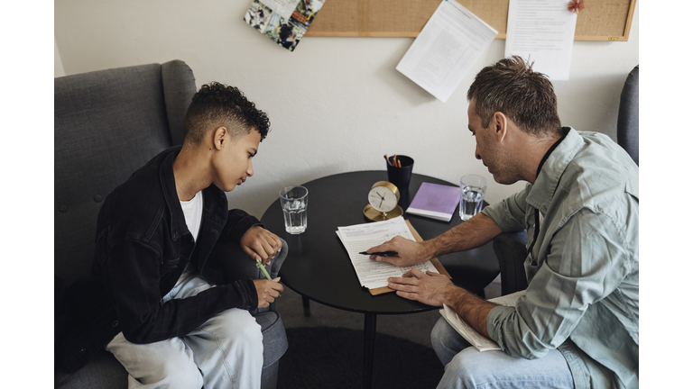 Male counselor discussing checklist with teenage student sitting at table in school office