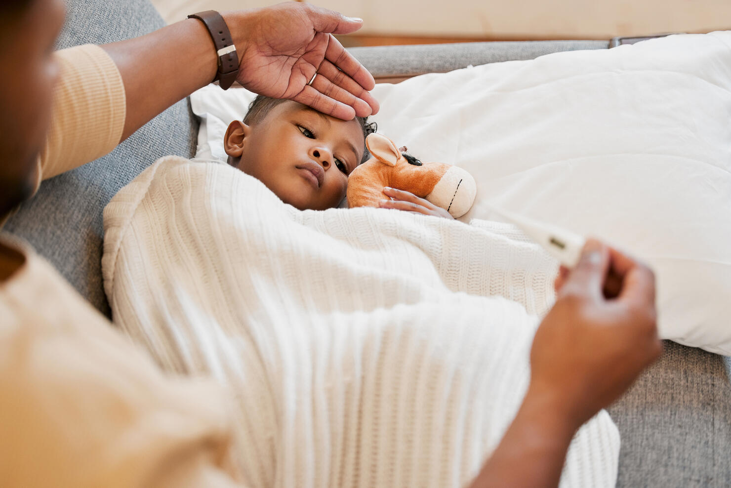 Sick, flu and cold child in bed, sad and ill with allergy and unwell at home with his worried mother. Parents hand on son's forehead for his temperature as she checks his fever with a thermometer