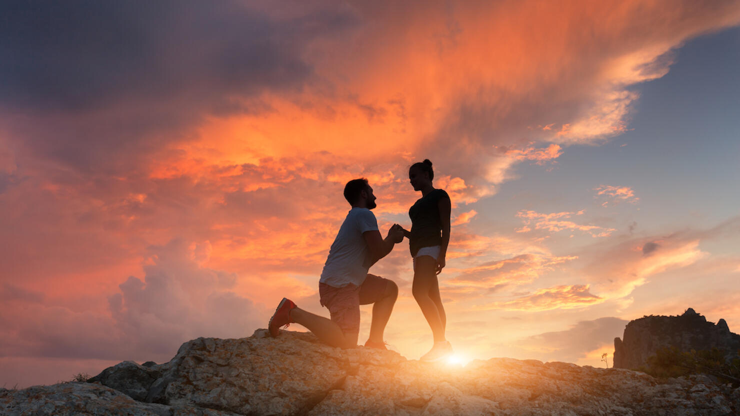 Silhouettes of a man making marriage proposal to his girlfriend on the mountain peak at sunset. Landscape with silhouette of lovers against colorful sky. Couple. People, relationship. Traveling couple