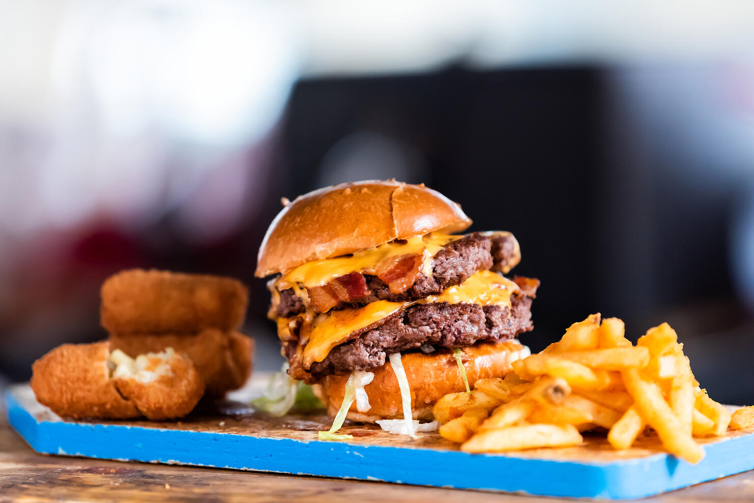 Burger greasy stacked street food, fried french fries closeup, melted cheese and bun on blue tray display in restaurant closeup, breaded fish sticks, bacon