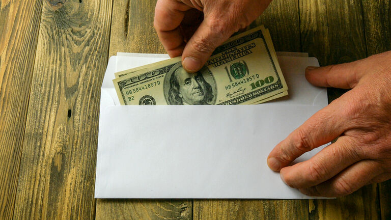 Human hands put two one hundred dollar bills in cash into white paper envelope that lies on brown wooden table. Concept of saving money or corruption and shadow economy.