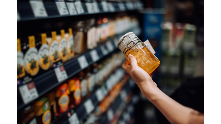 Cropped shot of young woman grocery shopping in supermarket, picking up a bottle of organic honey from the product aisle and reading the nutrition label on the bottle. Routine shopping. Healthy eating lifestyle