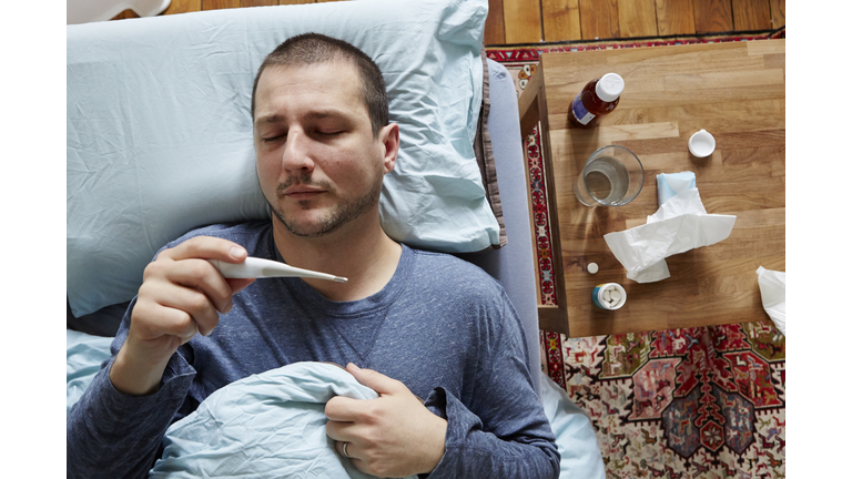 Mid adult man lying in bed looking at thermometer reading