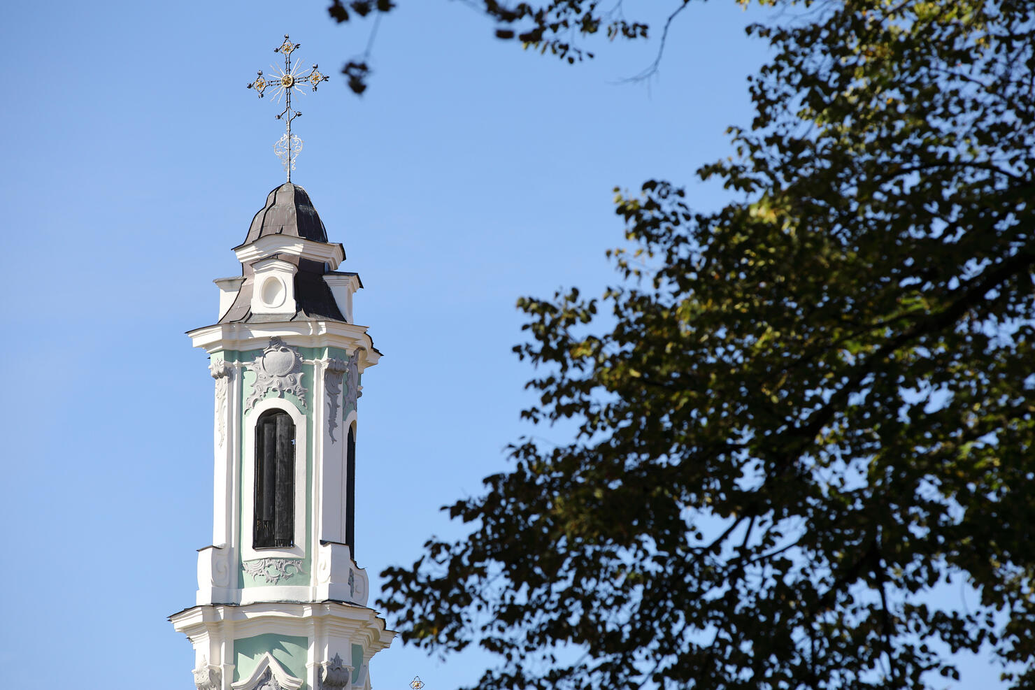 Church spire tower with cross against blue sky with tree in foreground