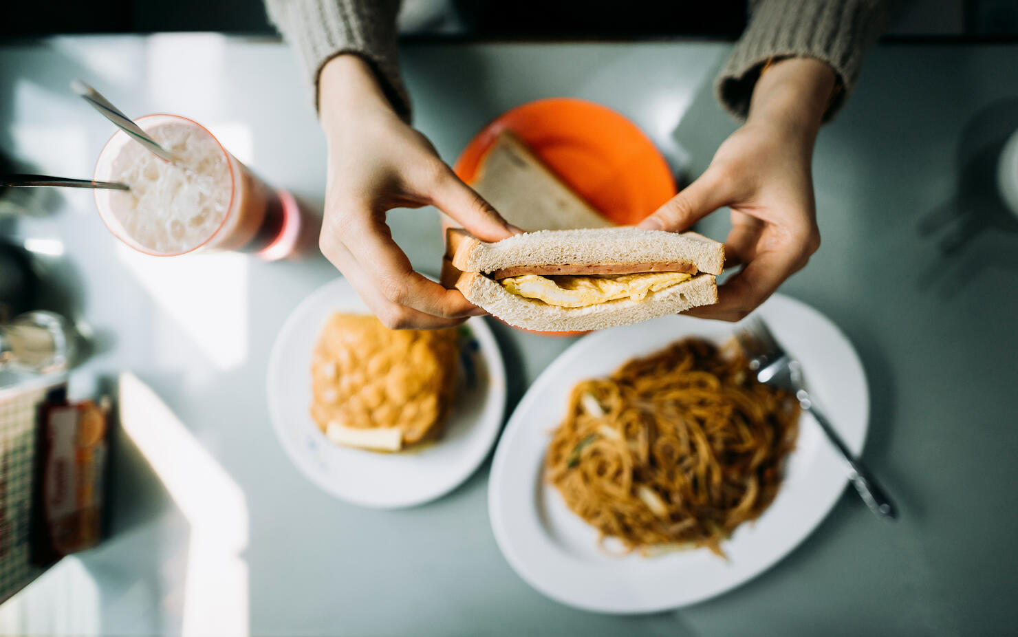 Overhead view of woman's hand holding Hong Kong style local foods, sandwich, buttered pineapple bun, fried noodles and iced red bean drink in traditional restaurant