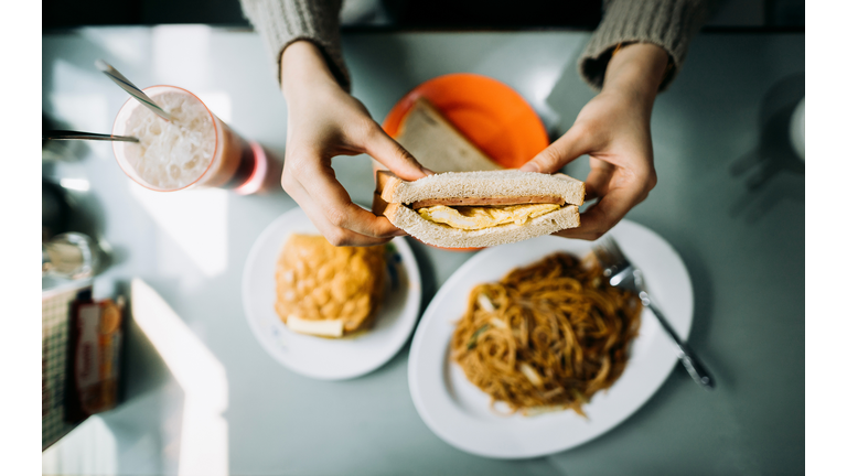 Overhead view of woman's hand holding Hong Kong style local foods, sandwich, buttered pineapple bun, fried noodles and iced red bean drink in traditional restaurant
