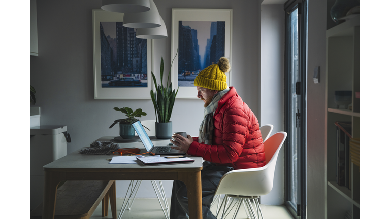 Mature man sitting working from home in a red puffer coat, scarf and wooly hat - Cost of Living Crisis