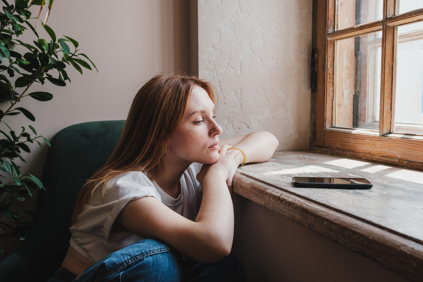 Upset redhead teen girl sitting by window looking at phone waiting call or message