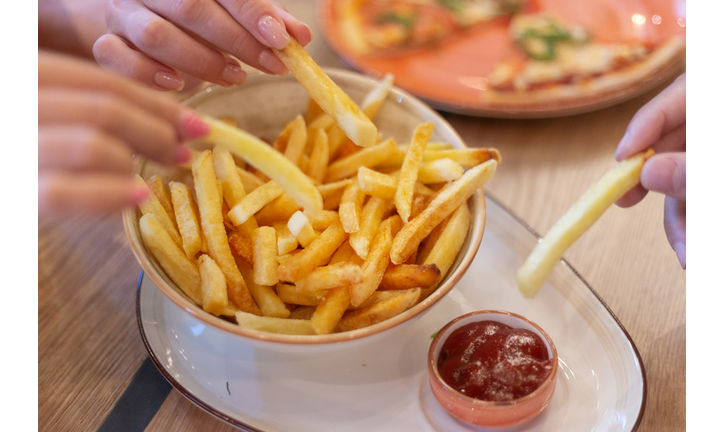 Hands of three young people sharing bowl of french fries and dipping them into ketсhup before eating in restaurant