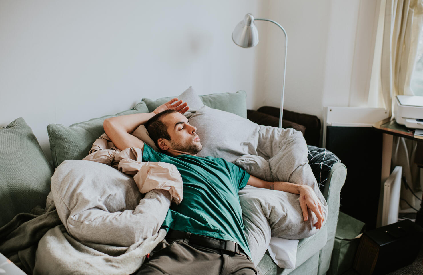 A man is asleep on a sofa surrounded by duvets and pillows