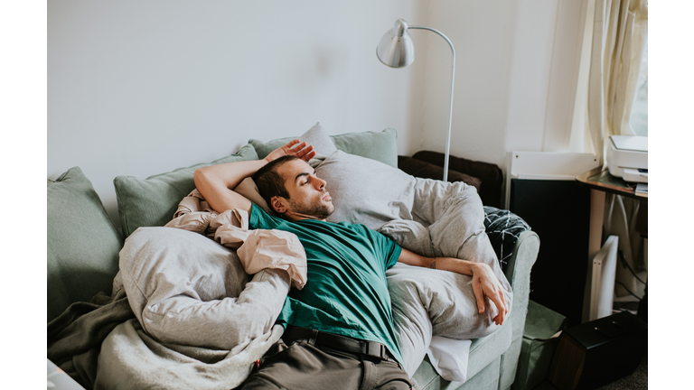 A man is asleep on a sofa surrounded by duvets and pillows