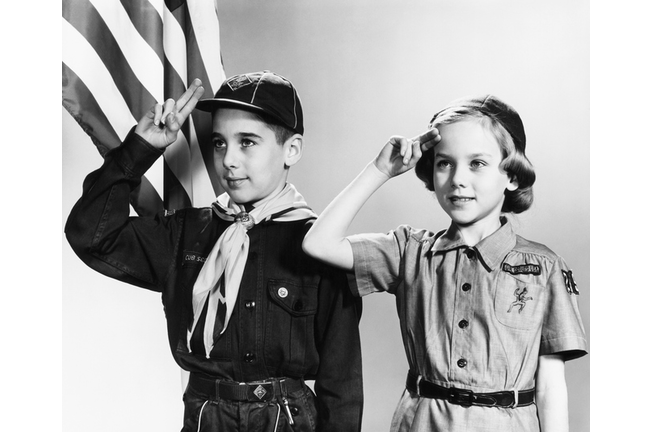 Boy and girl scouts saluting, American flag in background.