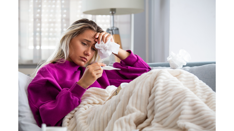 Sickness, seasonal virus problem concept. Woman being sick having flu lying on sofa looking at temperature on thermometer. Sick woman lying in bed with high fever. Cold flu and migraine.