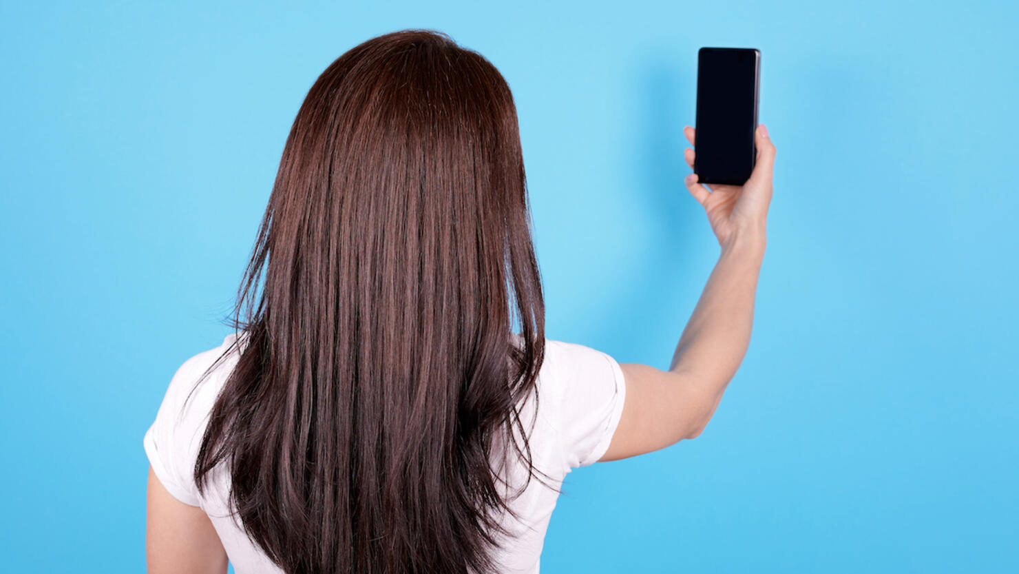 Brunette girl with long hair using mobile phone, make selfie, view from behind. Isolated on blue background.