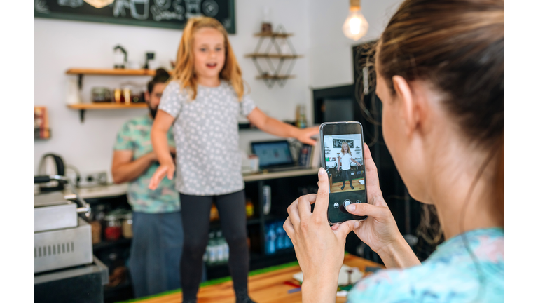 Mother recording her daughter dancing with the mobile while working in a coffee shop