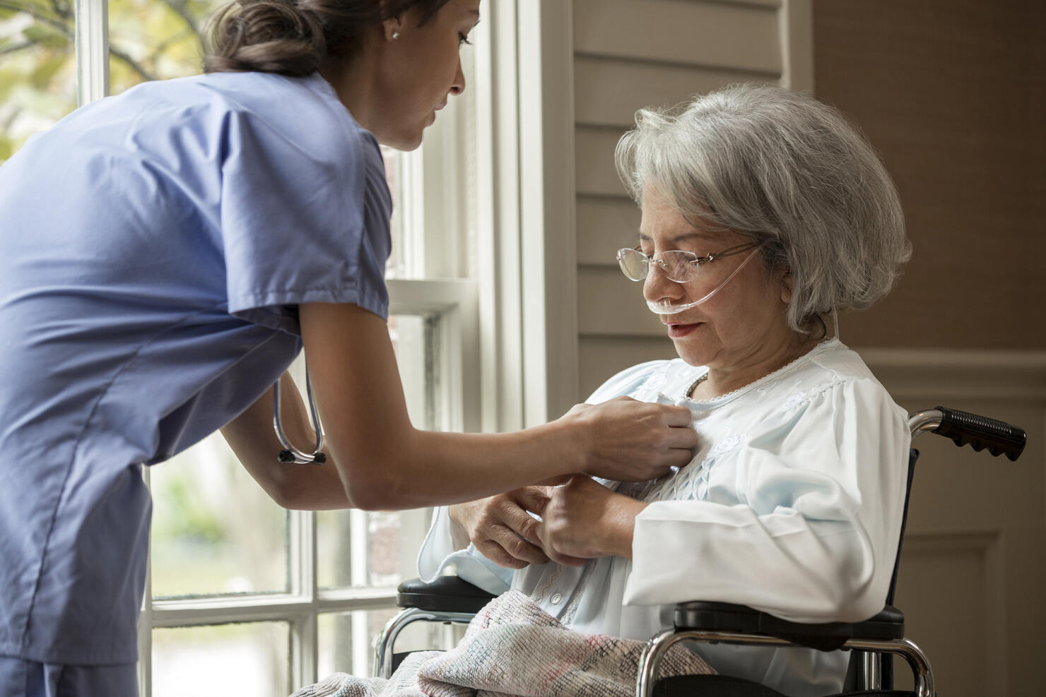 Nurse buttoning shirt of patient in wheelchair near window