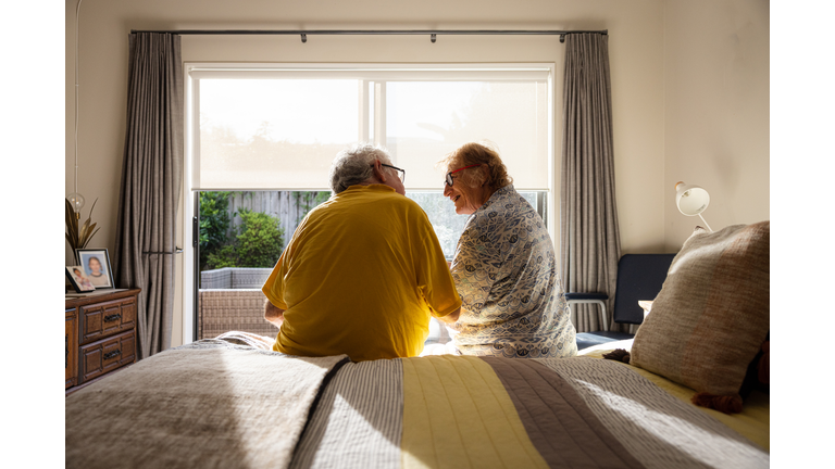Back shot of senior couple sitting on edge of bed, in front of window