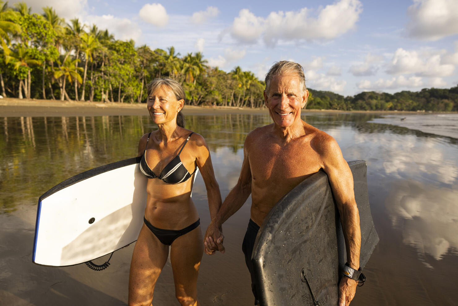 A senior couple holding boogy boards ready for a fun day in the ocean.