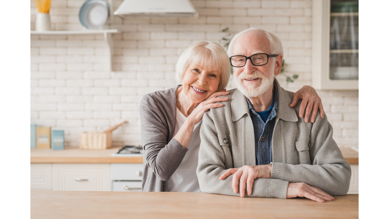 Portrait of caucasian smiling senior old elderly couple family spouses grandparents looking at camera, embracing hugging with love and care at home kitchen