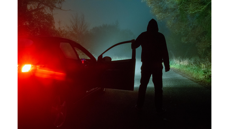 A spooky, mysterious hooded figure, standing next to a car with the door open. Looking down a moody, foggy, road at night