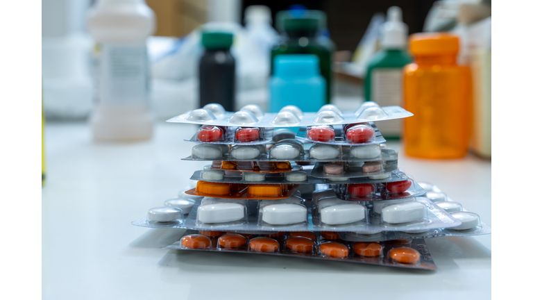The image depicts a neatly organized stack of blister packs containing various medications, including pills and capsules of different colors white, red, and orange