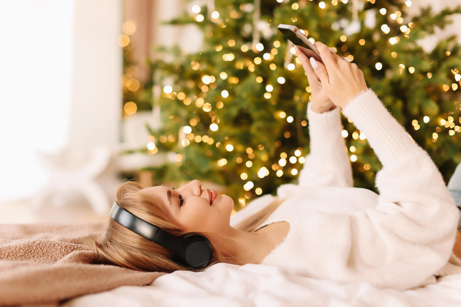 The girl is lying on the bed listening to music with headphones and looking into her mobile phone typing online messages in a decorated interior with a Christmas tree in the bedroom in December at home