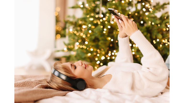 The girl is lying on the bed listening to music with headphones and looking into her mobile phone typing online messages in a decorated interior with a Christmas tree in the bedroom in December at home