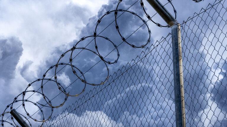 Close-up of metal fence with barbed wire over a stormy blue sky.