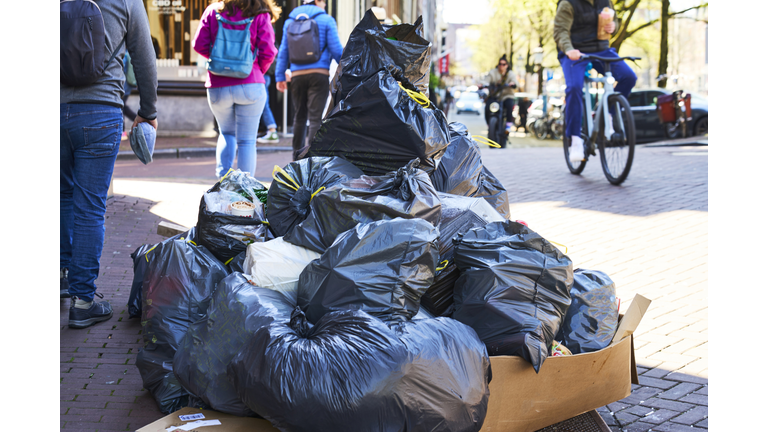 pile of garbage bags placed on city street curbside with people walking in the background, front view