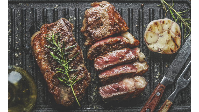 Close up of sliced roasted medium rare barbecue steak with rosemary, roasted garlic and cutlery on rustic iron grill