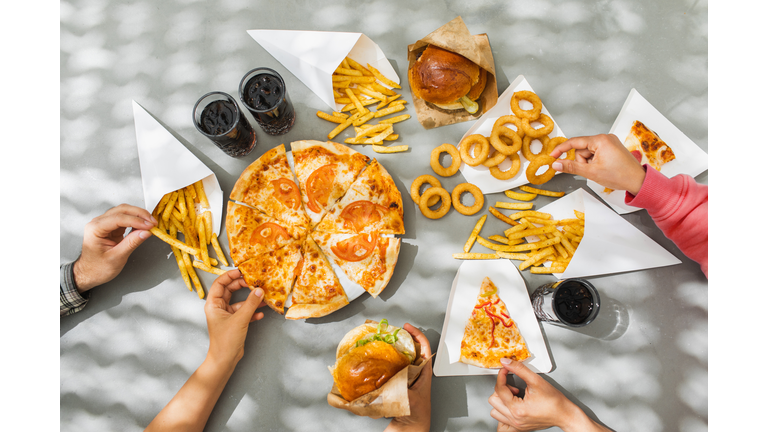 Human hands with assorted take out food such as pizza, french fries, onion rings, burger and cola.