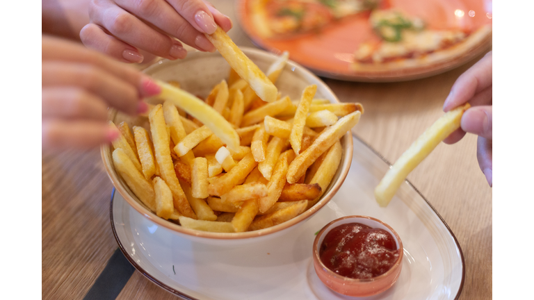 Hands of three young people sharing bowl of french fries and dipping them into ketсhup before eating in restaurant