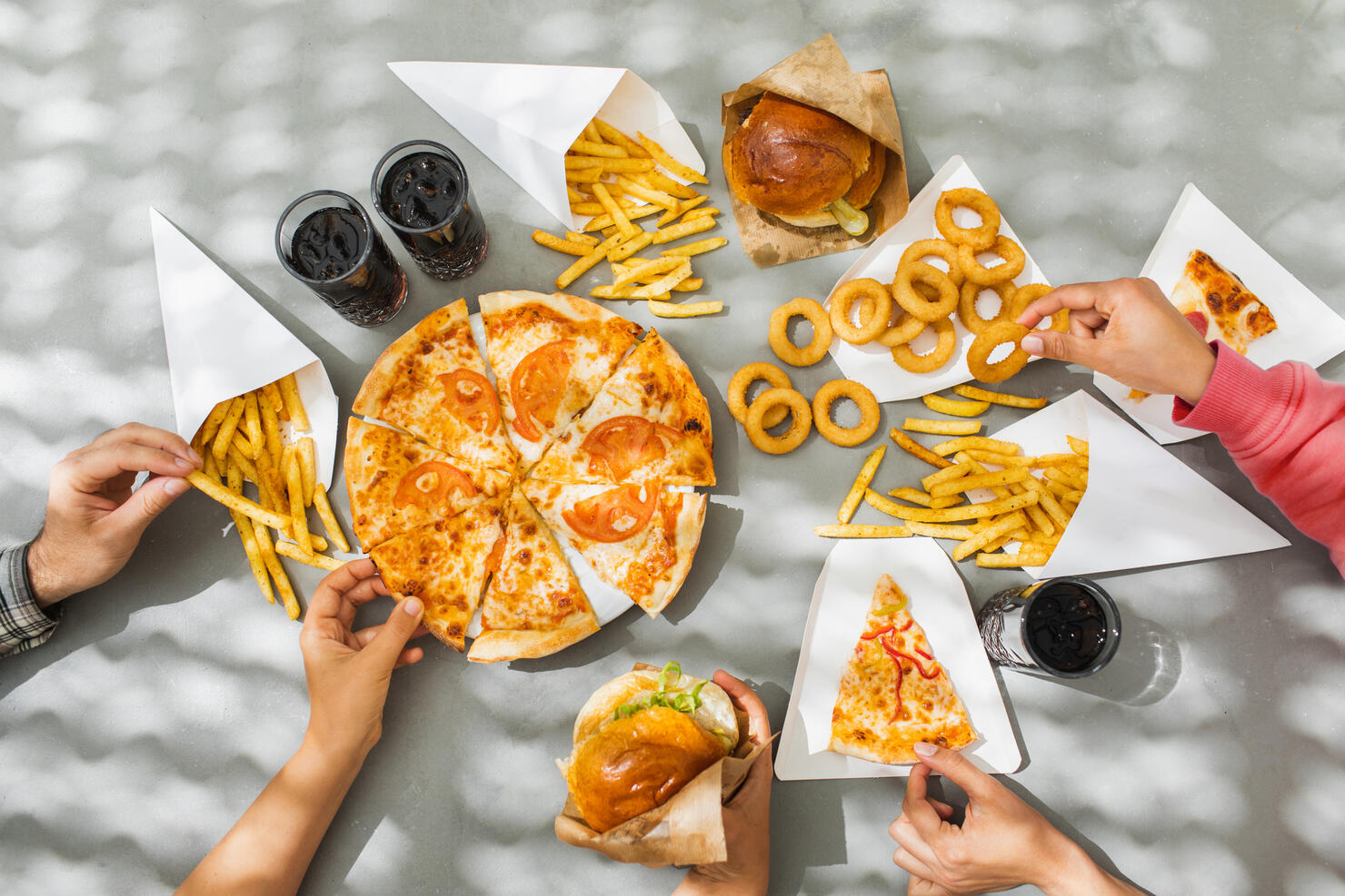 Human hands with assorted take out food such as pizza, french fries, onion rings, burger and cola.