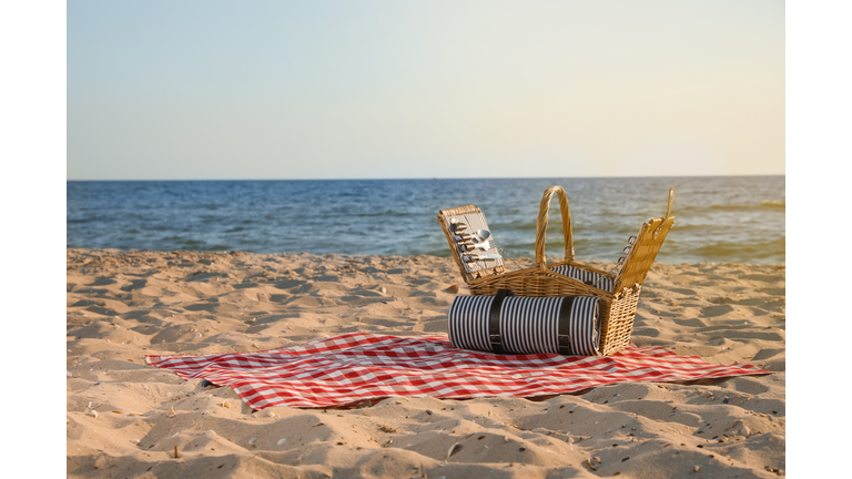 Blanket with picnic basket on sandy beach near sea, space for text