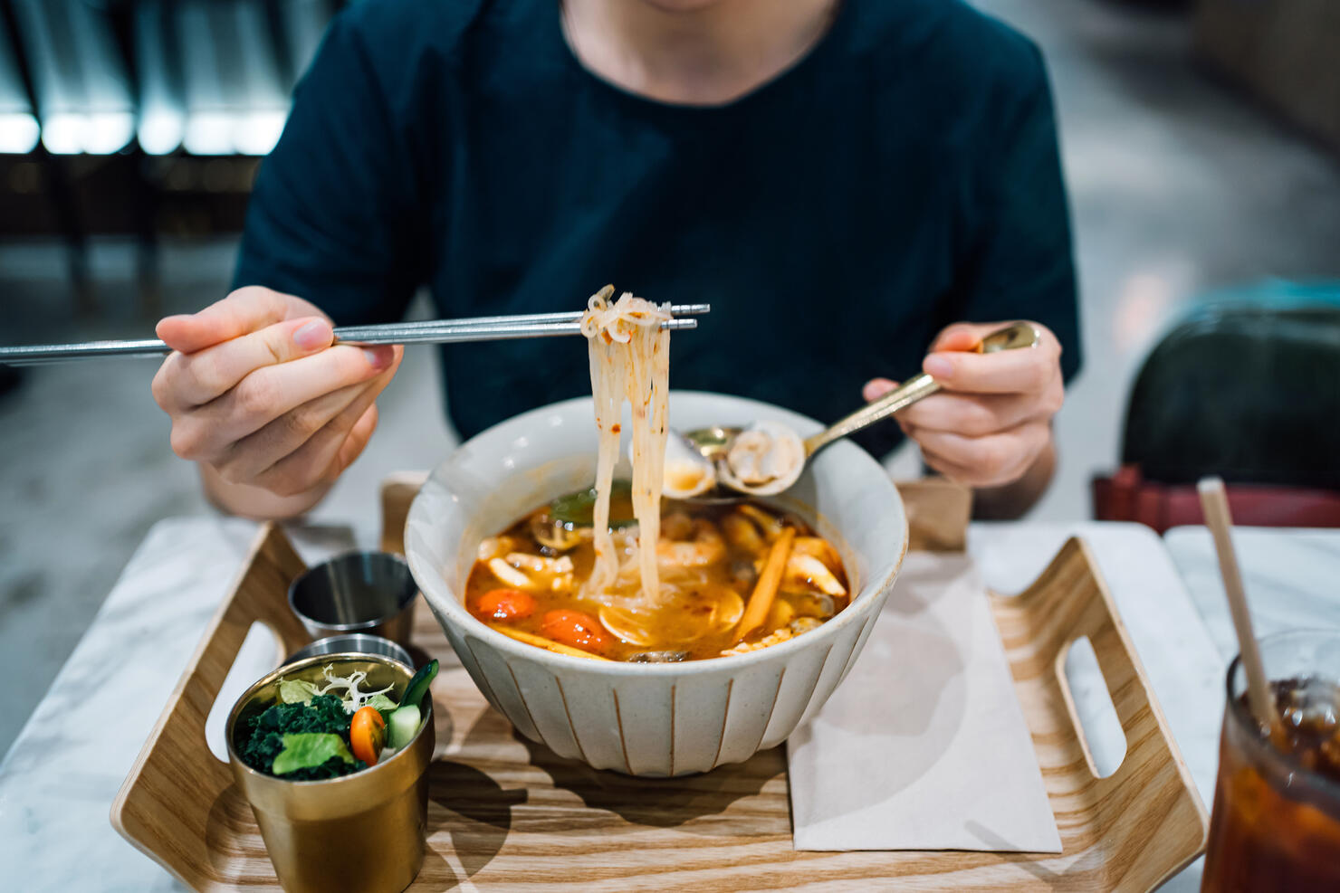 Close up of young Asian woman eating freshly served authentic Thai food, seafood Tom Yum Goong noodles (spicy prawn soup with seafood and noodles). With a refreshing and healthy green salad and iced tea in a Thai restaurant. An iconic taste of local Thai