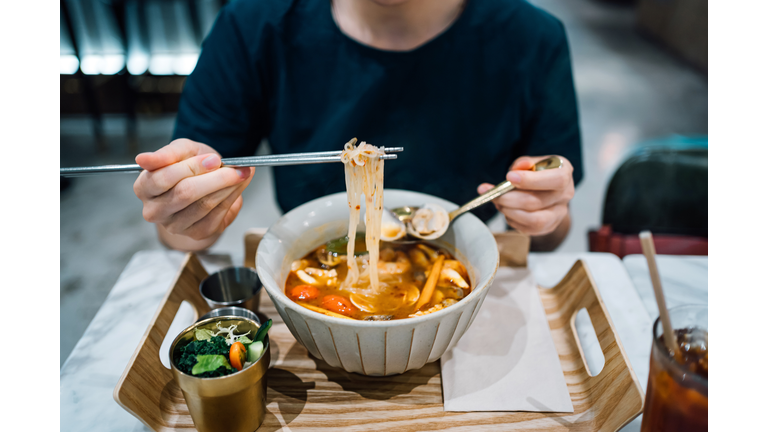 Close up of young Asian woman eating freshly served authentic Thai food, seafood Tom Yum Goong noodles (spicy prawn soup with seafood and noodles). With a refreshing and healthy green salad and iced tea in a Thai restaurant. An iconic taste of local Thai