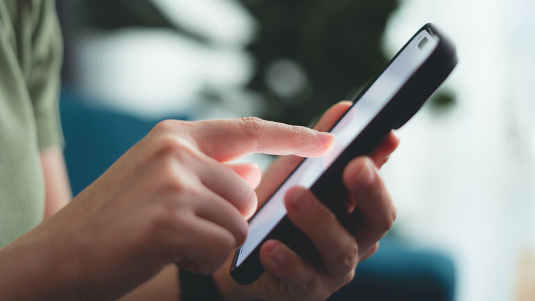 Close-up shot of female hands using smart phone sitting on sofa in living room at home.