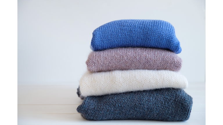 Knitted woolen things, stacked, lie on a white wooden table or shelf. Warm sweaters for charity. The concept of storage, care and washing of handmade products. A copy of the text space. Thrift store, assistance to migrants and refugees. Secondary use.